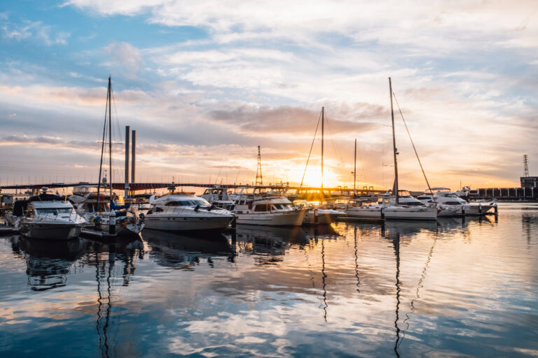 sunset on a marina with several yachts and motor boats moored in the foreground and a large bridge in the background